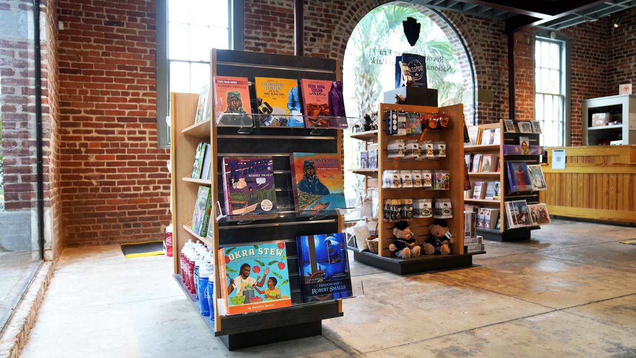 A small bookstore with three shelves of books and trinkets laid out in front of a three windows and a desk.