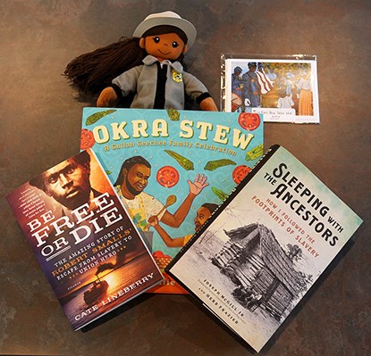 Three books "Be Free or Die", "Okra Stew", "Sleeping with the Ancestors", sitting on top of a Black female ranger doll, and a postcard of a painting of Black soldiers holding a flag.