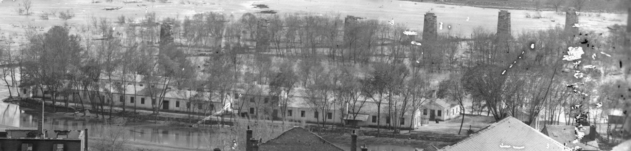 A black and white photograph of numerous small, white buildings on an island in a river.