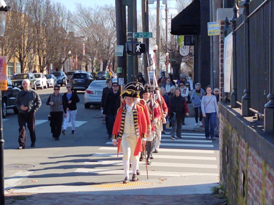 Costumed reenactors in red Revolutionary War uniforms march in a line down a modern city street.