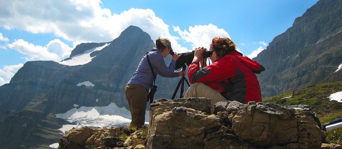 Two women scan mountainside for mountain goats; one looks through scope, the other through binoculars.