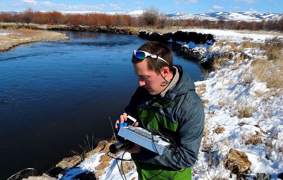 Jerry O'Neal research fellow Andrew Spencer stands by river holding monitoring equipment.