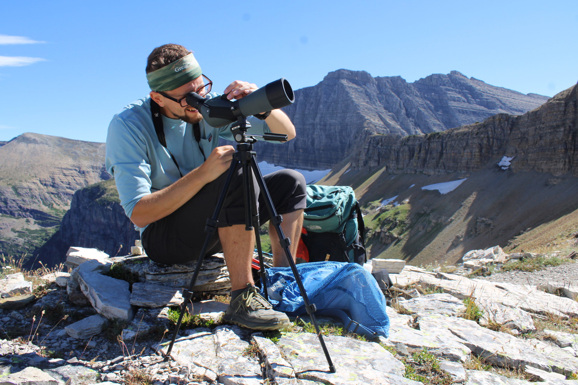 A man looks through a scope while sitting on top of a mountain.