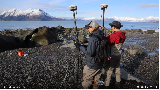 Still-frame of a video; two researchers holding monitoring equipment stand on a rocky shoreline.