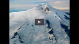 Still-frame of a video; aerial view of a snow-covered, pointed volcano.