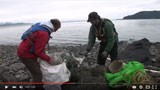 Still-frame of a video; two people pick up trash on a beach.