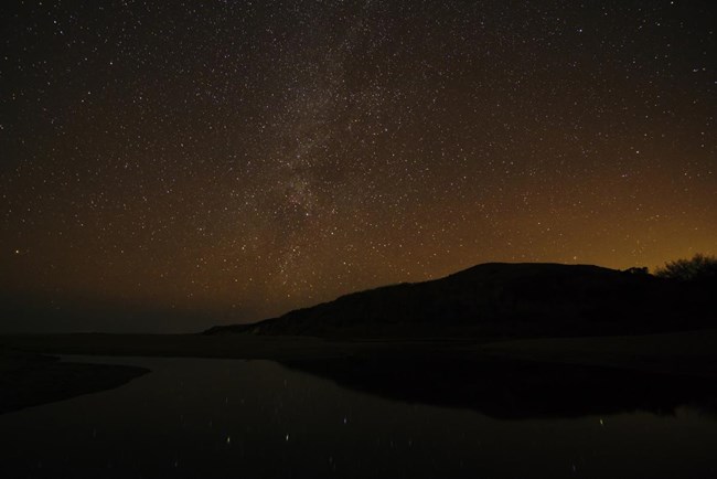 The Milky Way visible over a creek flowing across the beach and into the ocean, with light pollution also obvious on the horizon