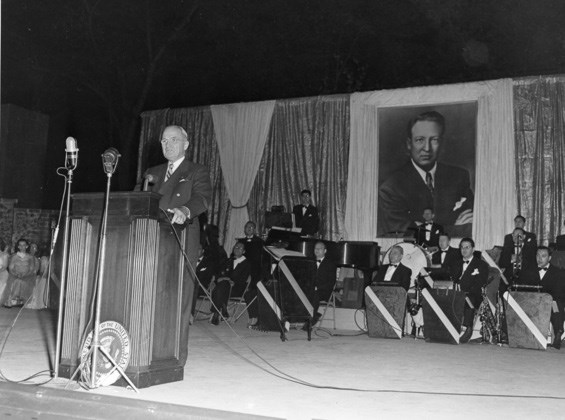 President Truman speaks at a podium with a large portrait of Carter Barron behind him.