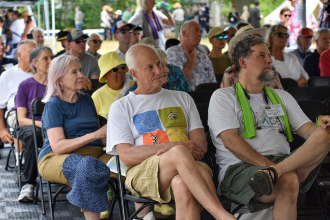 A small audience of people are seated and listening to a presenter out of frame to the right. They are all under an outdoor tent.