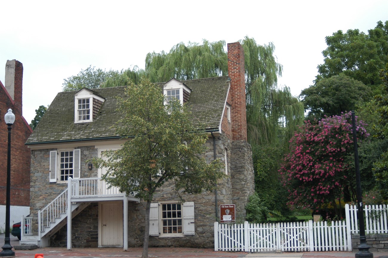 Two and a half story stone house with adjoining green space lot is visible behind a small tree.
