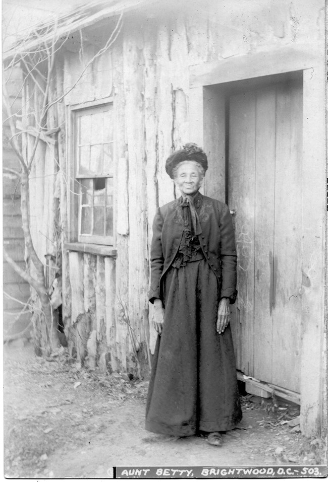 Woman wearing a heavy, long black dress and jacket and hat stands next to a log structure. Elizabeth Proctor Thomas, African American woman who owned the property where Fort Stevens was constructed during the Civil War.