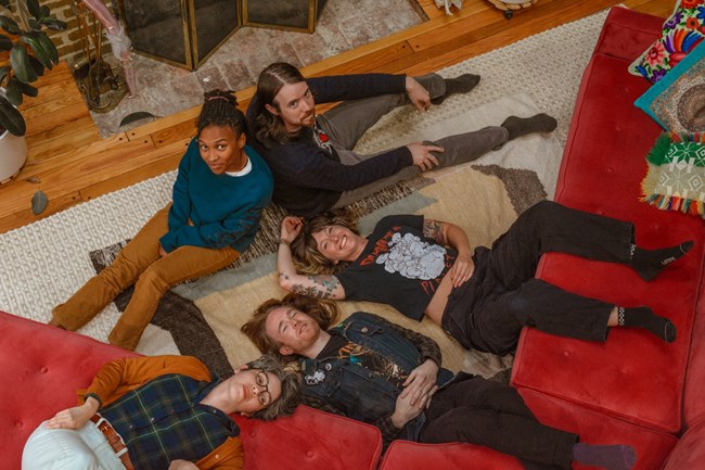 Five people lounge on the floor by a red couch smiling.