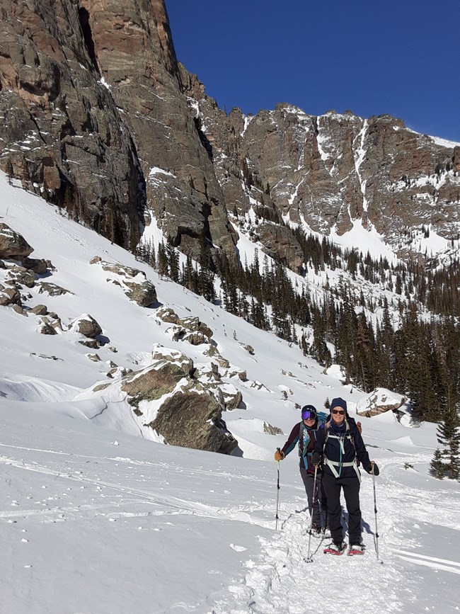 Two people are snowshoeing above Loch Vale on a clear, sunny day