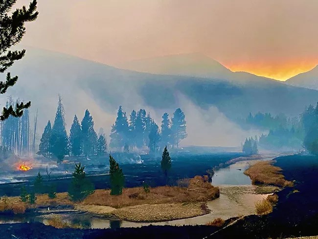 A river flows through burned meadows and forested areas, with smoky skies behind and a glimmer of a colorful sunset.
