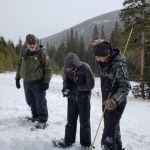 Two students with science tools stand with a park ranger on a snowy landscape near a forest.