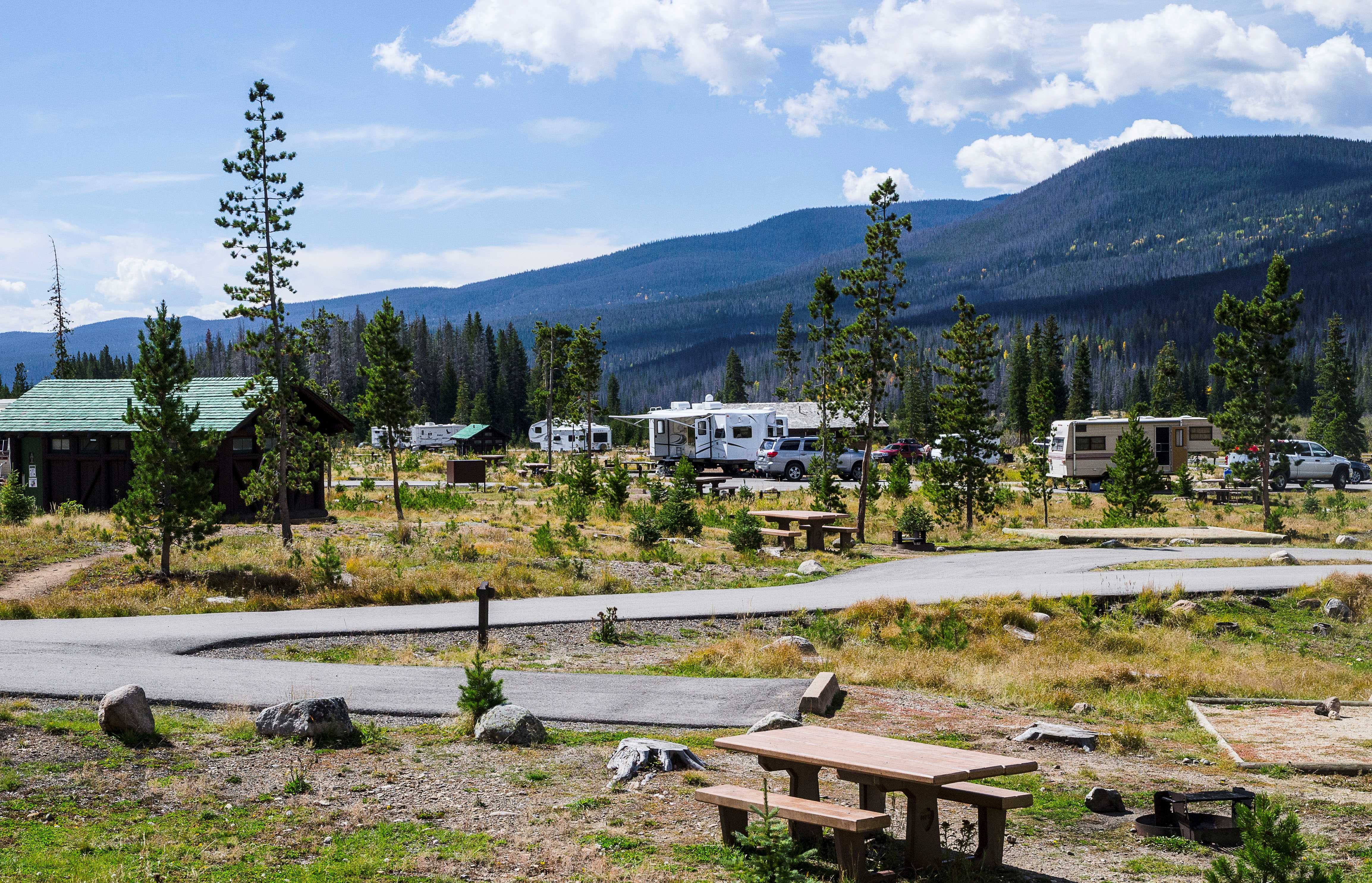 Tents and RVs are set up in campsites in Timber Creek Campground on a sunny day in summer. The campground has a paved road and is lined with pine trees. There is a restroom building in the middle of several campsites.