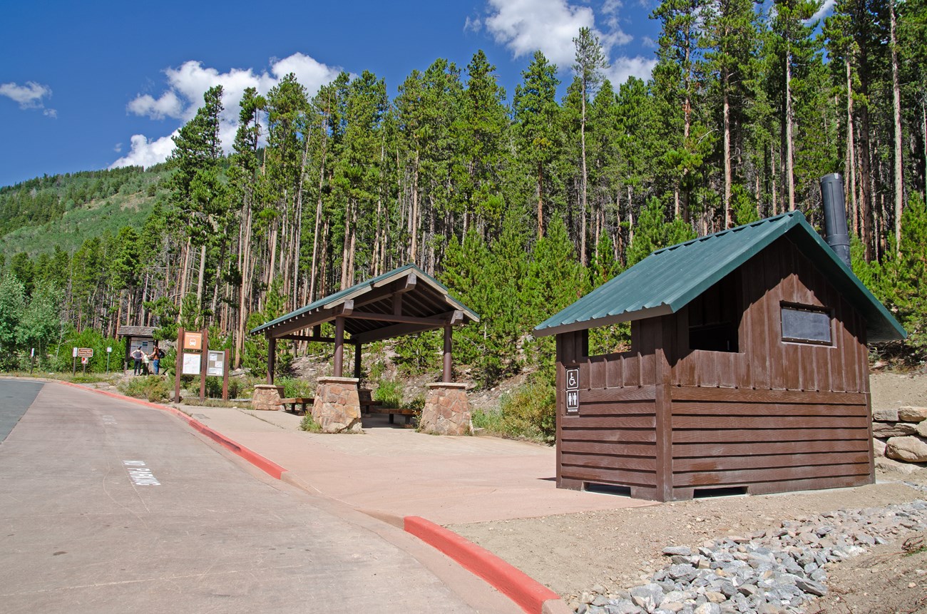 A new vault toilet building has been constructed near the Bierstadt Lake Shuttle Stop and Bierstadt Lake Trailhead. The new building is dark brown with a dark green roof. There are tall pine trees in the background.