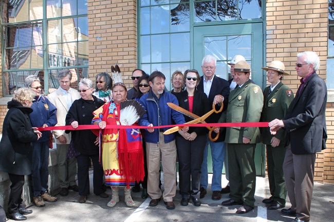 A ribbon-cutting ceremony in front of a brick building with large windows. A diverse group of people, including National Park Service rangers in uniform, community members, and representatives, gather to celebrate. A person in traditional attire holds a