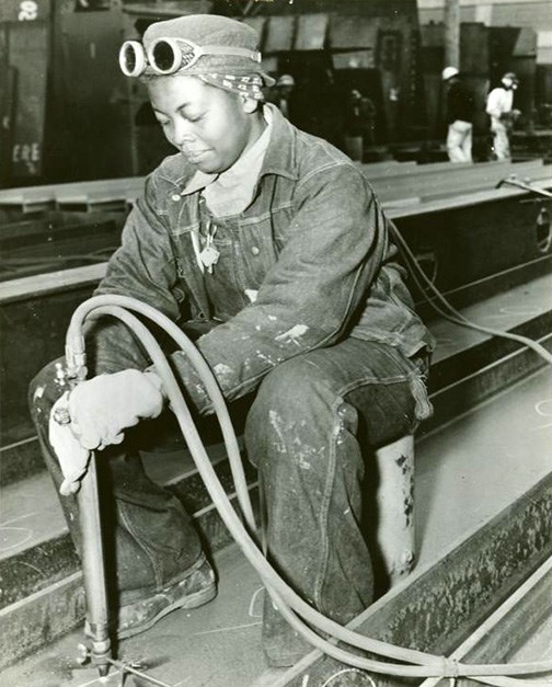 An African American woman in work clothes is working on a ship. She is kneeling and holding a tool connected to hydraulics.