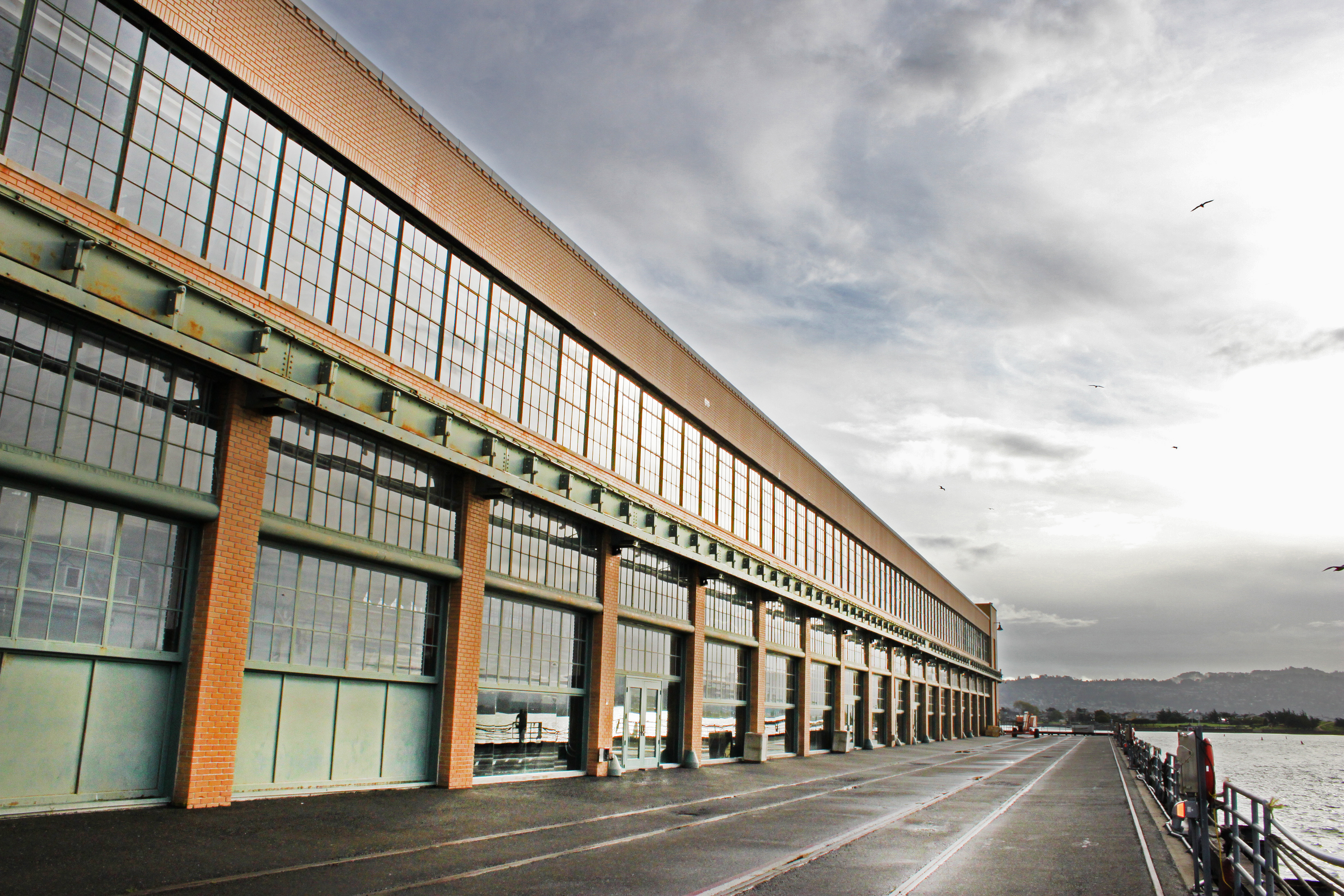 A long, rectangular industrial building with large, grid-patterned windows spans the length of a waterfront area. The building's facade is characterized by brick and steel construction, reflecting the architectural style of the early 20th century.