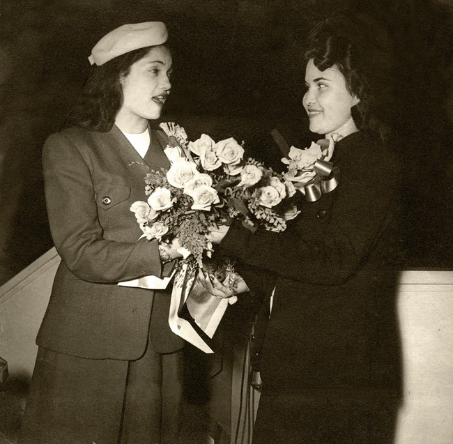 Two women in suits and hats are holding flowers.