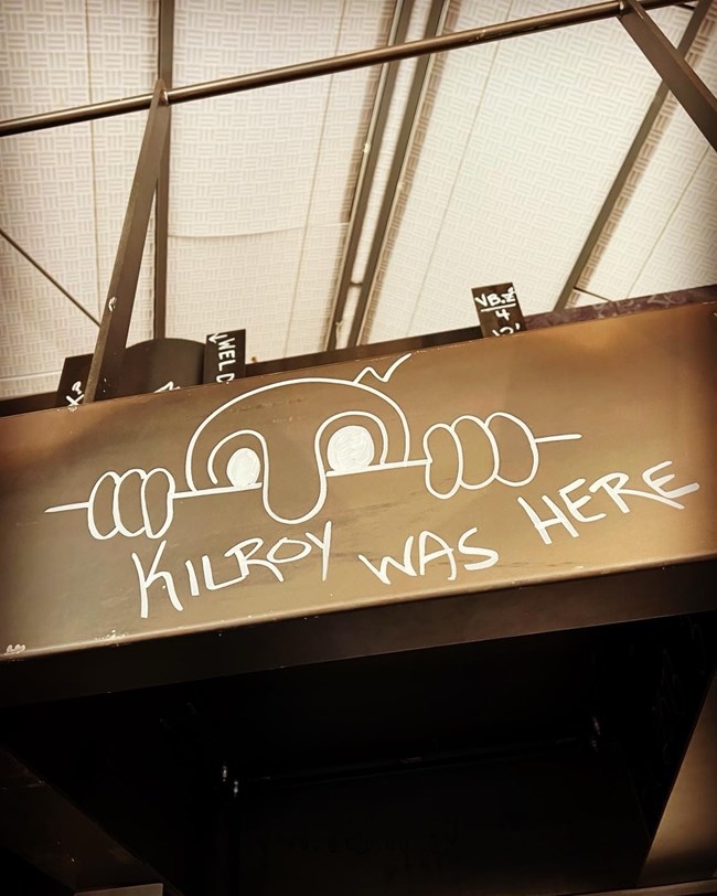 Photo of "Killroy was here". Painting of a bald male character looking over a ledge with its hands and nose hanging over.