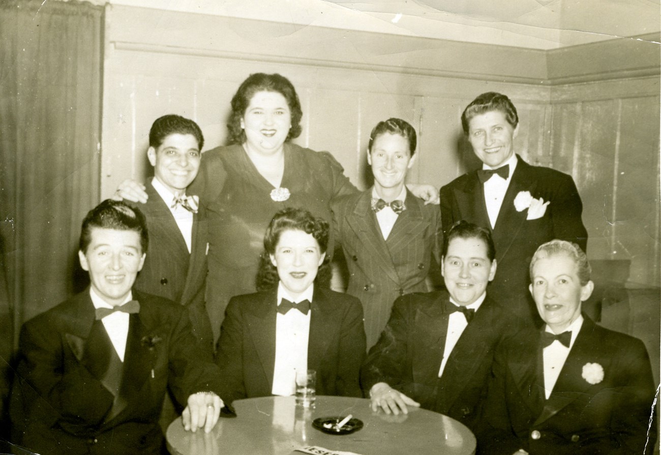 Male impersonators sit at a table in tuxedoes and fashion dress. Historic photo.