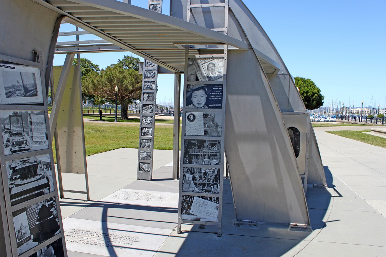 A metal structure which is part of the Rosie Memorial. Photos of women can be seen, along with the natural surroundings of a park.