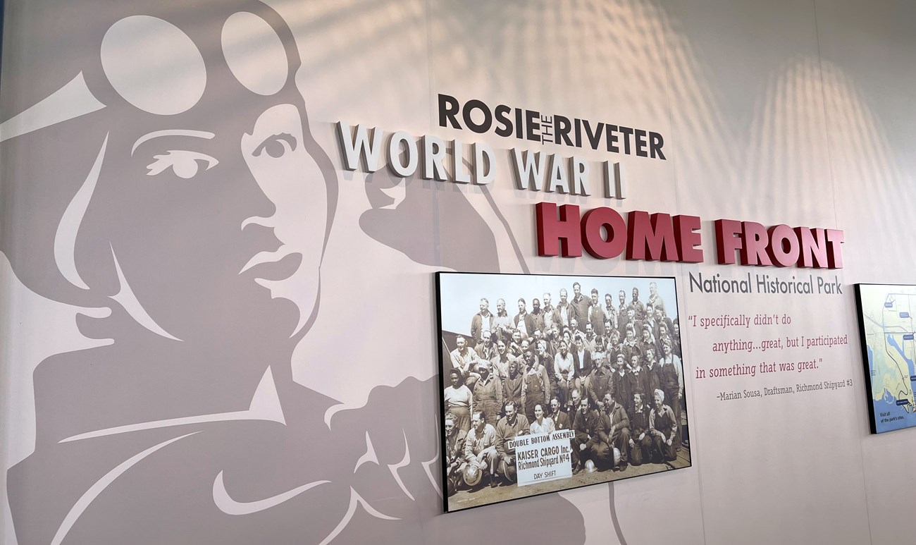 Entrance display at the Rosie the Riveter/World War II Home Front National Historical Park showing a large illustration of a female worker representing Rosie the Riveter, a historical group photo of shipyard workers, and a quote from Marian Sousa, a WWII