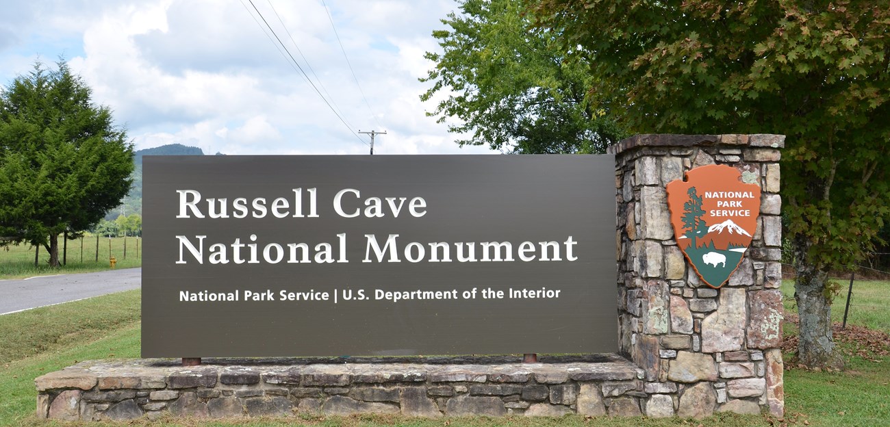 The entrance sign for Russell Cave outside, along roadway.  Sign says " Russell Cave/ National Monument/ National Park Service/ U.S. Department of the Interior".