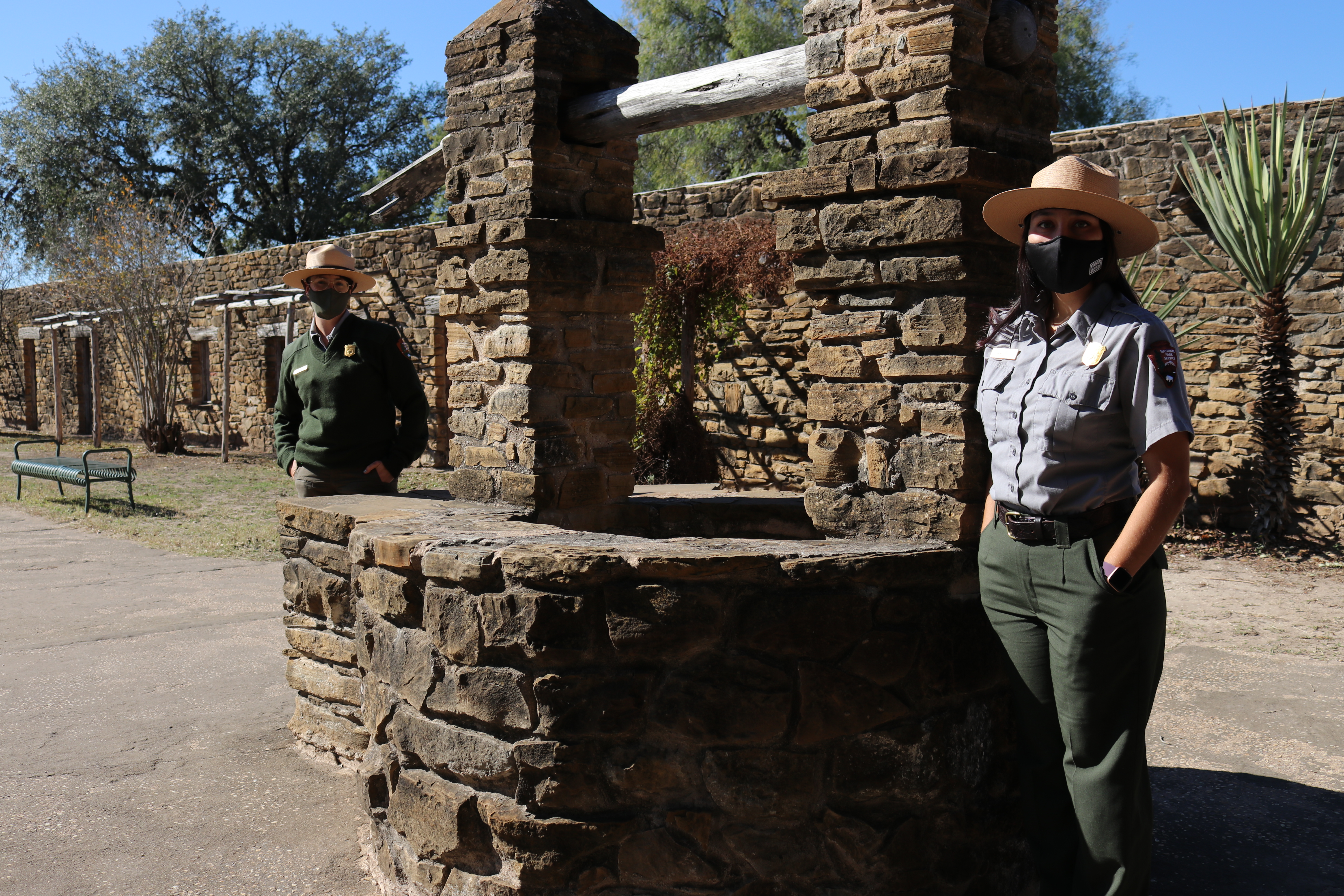 Two park rangers stand on opposite ends of the well at Mission San Jose wearing masks.