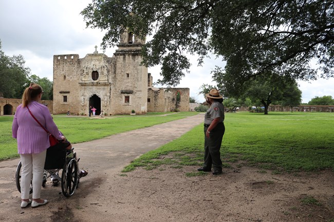 A ranger points toward Mission San Jose while a person pushes another person in a wheelchair.