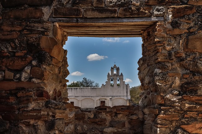 Looking through a window in a stone wall toward the bell tower of a mission.