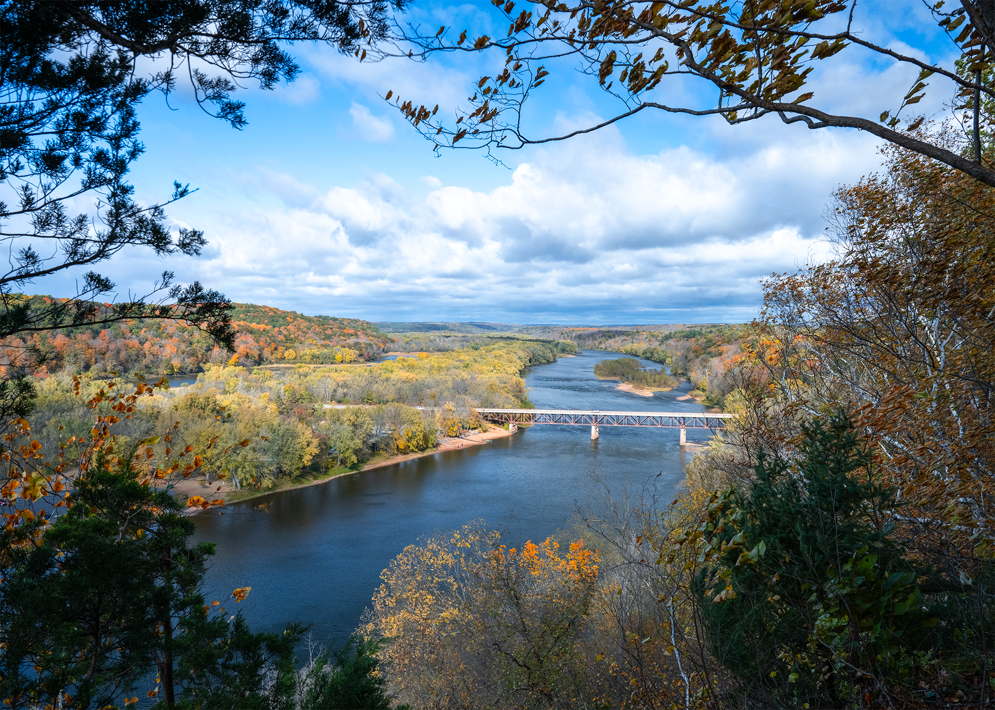 A river reflects a deep blue from a partially cloudy sky while winding through a forested landscape dotted with rich autumn colors. A bridge crosses the river in the midground and the scene is framed with dark leaves and branches.