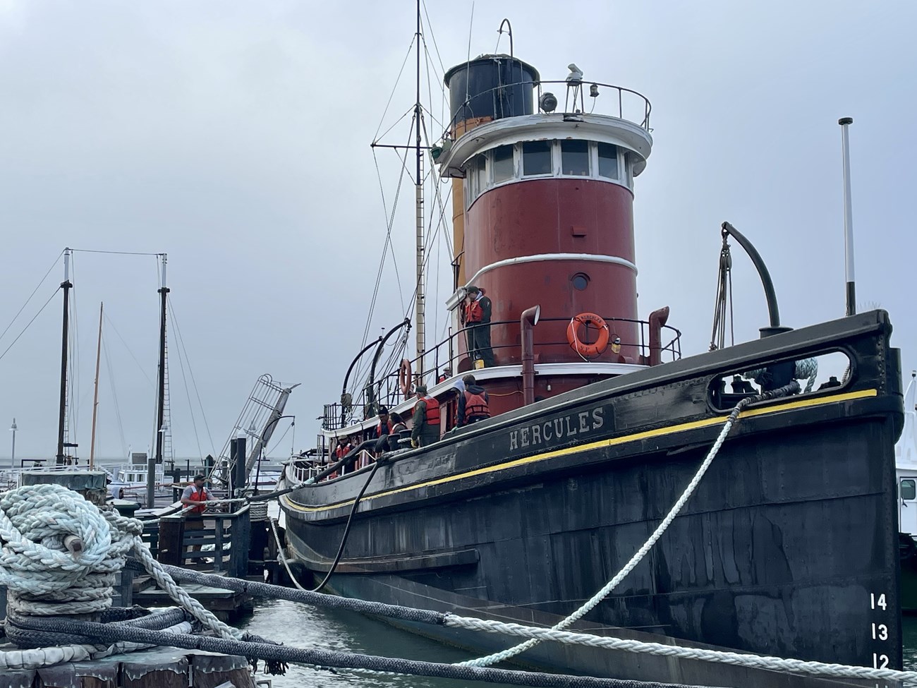 the steam-tug Hercules is moored to a dock while workers wearing life jackets work on deck