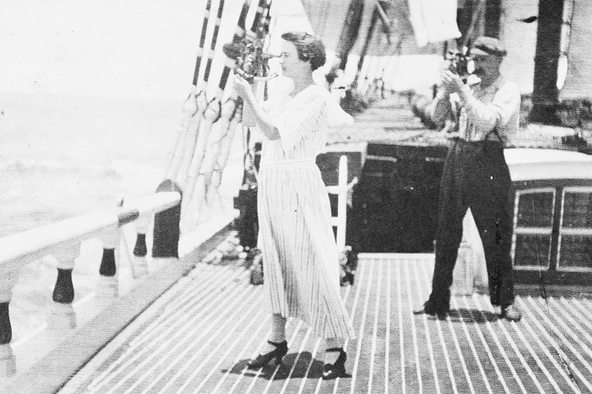 A woman in a white dress stands on the deck of a ship with a navigation tool held up to her eye