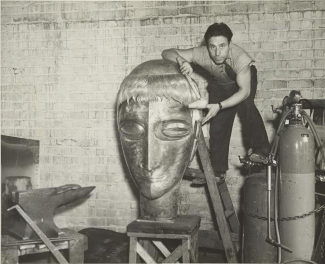 Beniamino Bufano looks at the camera while on a ladder, crouching over a large sculpted head