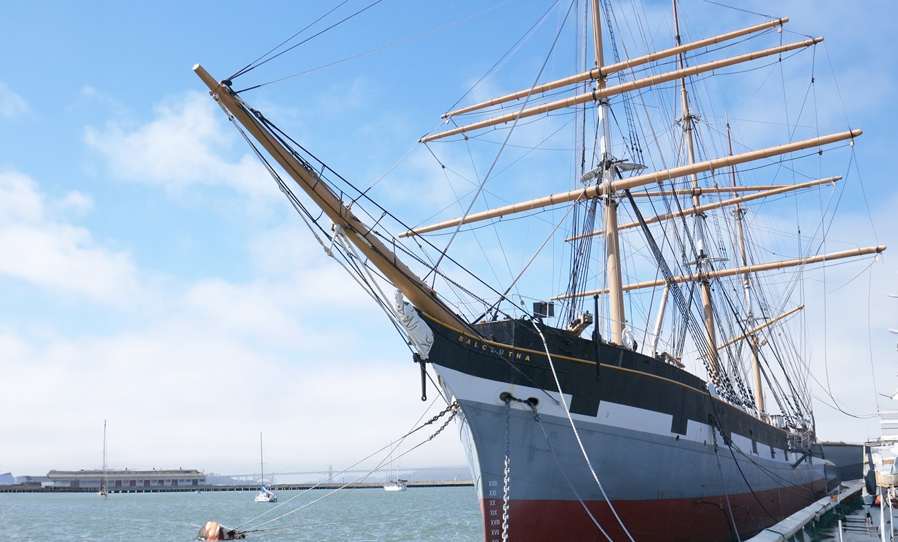 profile of the ship Balclutha, with tall wooden masts and a hull with red, black, and gray horizontal stripes