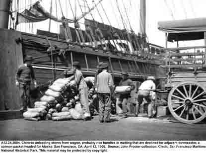 A black and white photo taken in 1906 of a San Francisco pier. Chinese men are unloading rice wrapped in matting from a cart. 