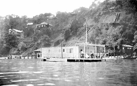 A black and white photo showing a flat-roofed houseboat moored in a cove with a hillside behind it.