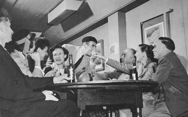 Black and white photo of several artists sitting around a table smiling and laughing together