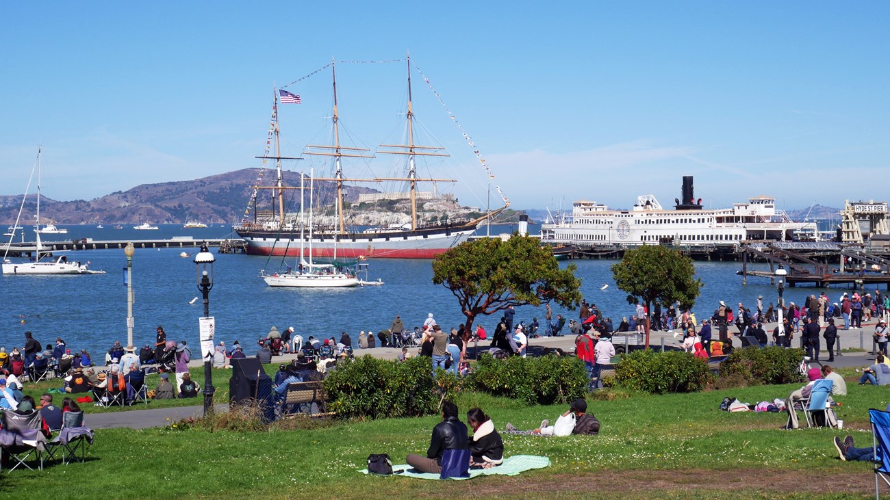 people sit on a grassy lawn and look out over Aquatic Park Cove with Balclutha in the background