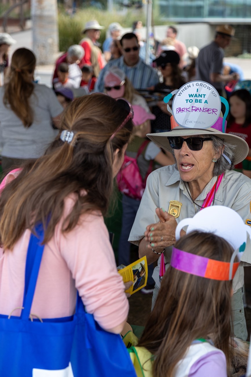 A female park volunteer stands on the far side of a table, with desert props, speaking to a mother and daughter on the close side of the table facing away from the camera. The volunteer is wearing brown pants, a tan button up, and a wide brimmed hat.