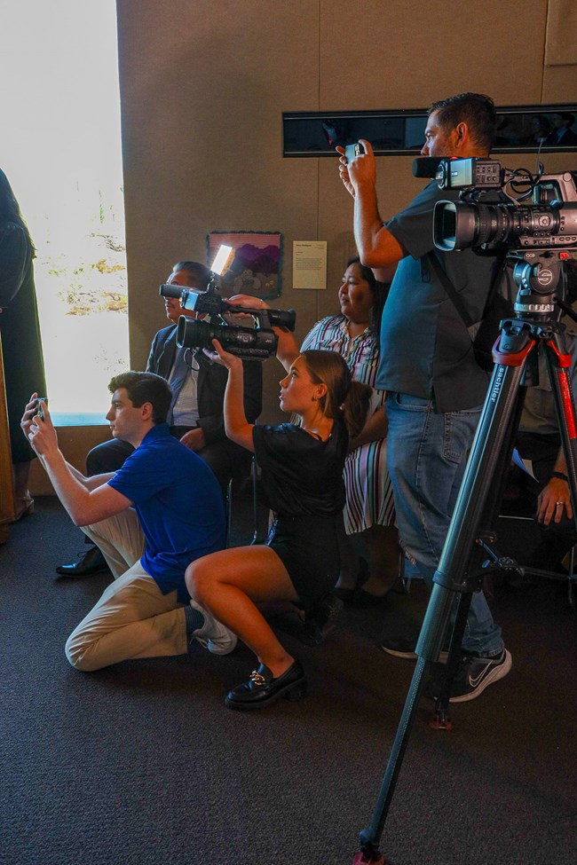 A group of reporters huddle closely together, some standing in the back while those in front kneel. All have a camera of some form in their hands, recording something to the left of the image.