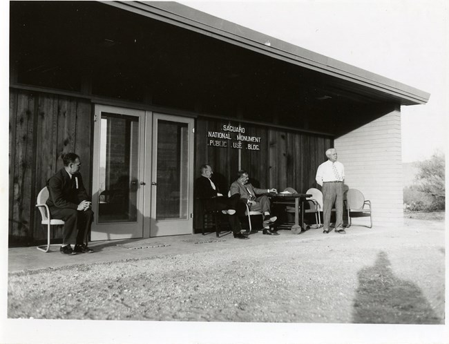 An old black & white photo shows part of the historic visitor center, with four older white men sitting around outside. The wall of the building reads "Saguaro National Monument" "Public Use BLDG"