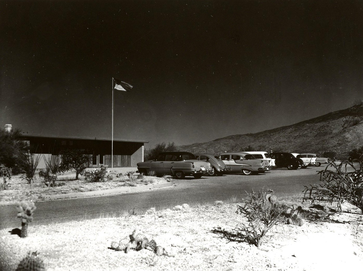 Old black & white photo shows a historic look at the Rincon Mountain Visitor Center, with 7 classic cars in the parking lot. Appearing to be from the late 60s.