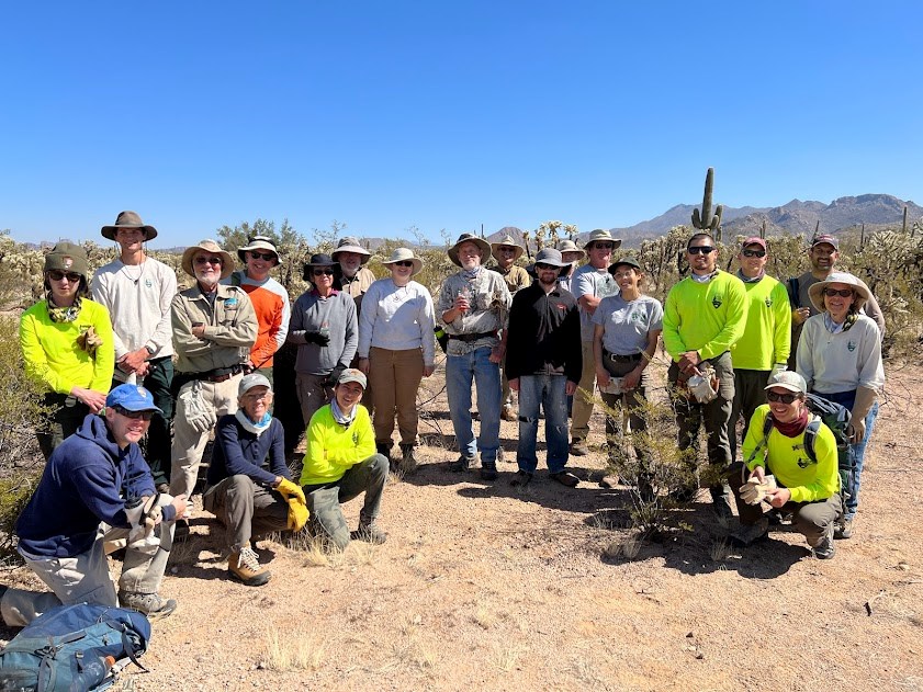 A large group of volunteers and park staff pose for a picture in the desert. They are wearing field clothing and safety gear and are smiling.