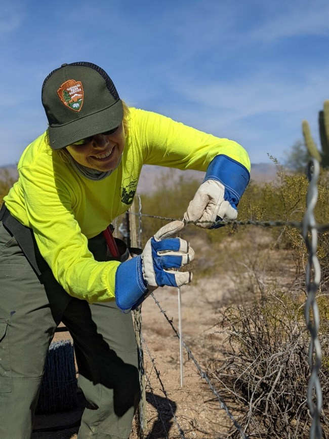 A crew member works on constructing fence line. They are wearing protective gear and smiling while adjusting fence wires.