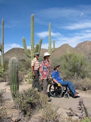 Three people, two walking one in a wheelchair, on a paved path in a desert setting.