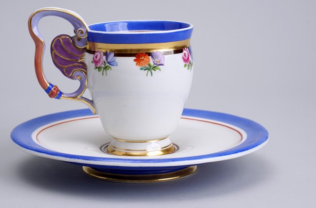 Hot chocolate cup and saucer with blue and gold trim.  Cup handle is in the shape of a butterfly and is colored purple and gold.  There are flowers painted on the cup.